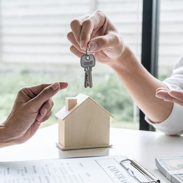 A person handing over house keys above a small model house on a table.